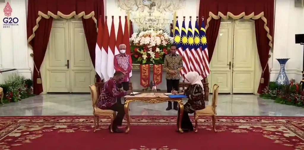 Ida Fauziyah and Dato’ Sri M. Saravanan duduk dan menandatangani Nota Kesepahaman Penempatan dan Perlindungan TKI Sektor Domestik bersama Presiden Joko Widodo dan Perdana Menteri Dato’ Sri Ismail Sabri Yaakob yang berdiri di depan bendera Indonesia dan Malaysia.