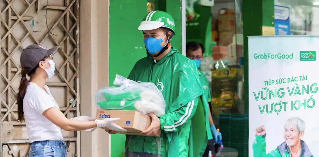seorang wanita menyerahkan satu set paket bantuan kepada seorang pria dalam seragam grab hijau dalam rangka program bantuan makanan.