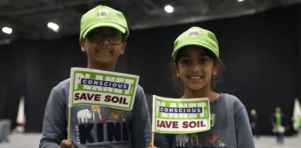 Dua anak dalam baju yang mirip memegang bendera berbunyi ‘Save Soil’