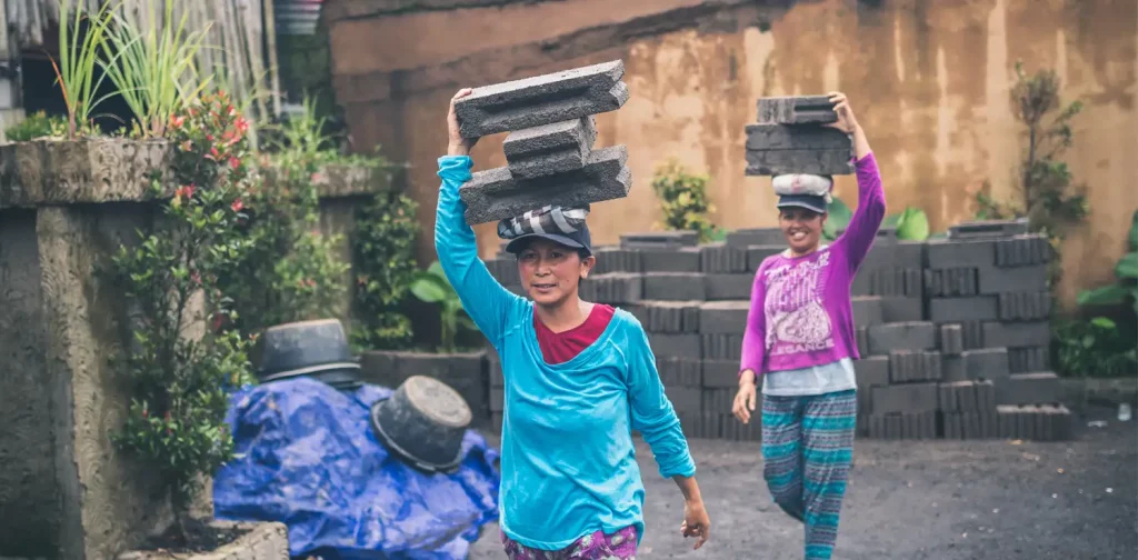 Dua wanita di Bali berjalan membawa batu bata beton di atas kepala.