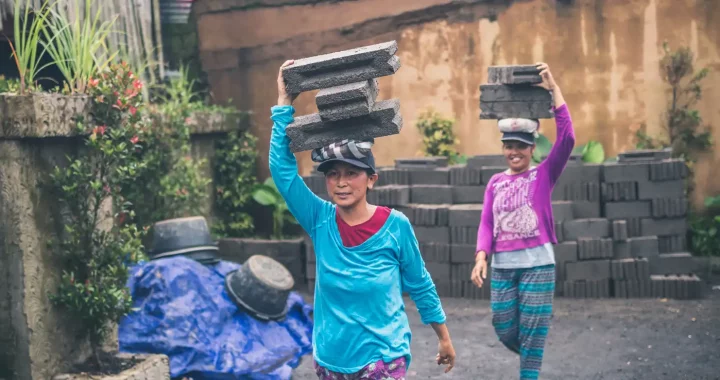 Dua wanita di Bali berjalan membawa batu bata beton di atas kepala.