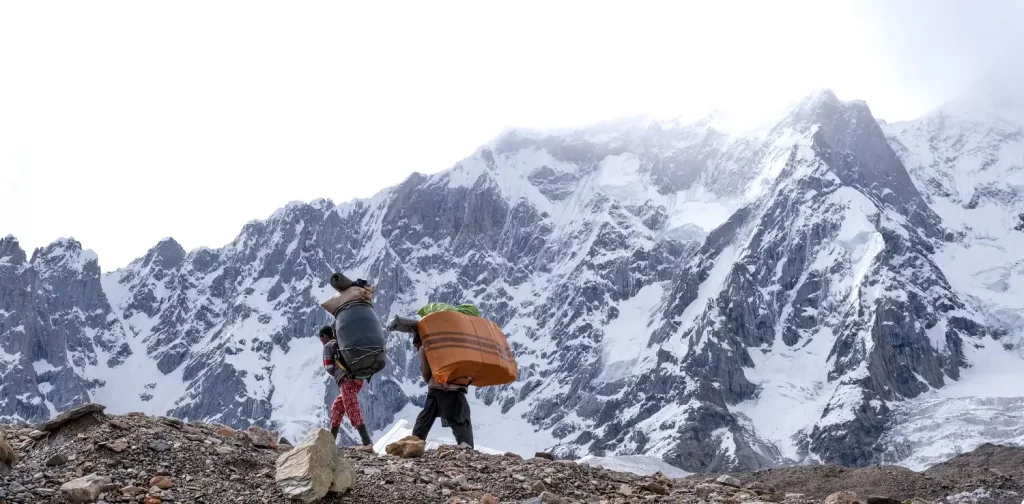 dua orang membawa barang di punggung mereka di gletser baltoro dengan latar gunung es