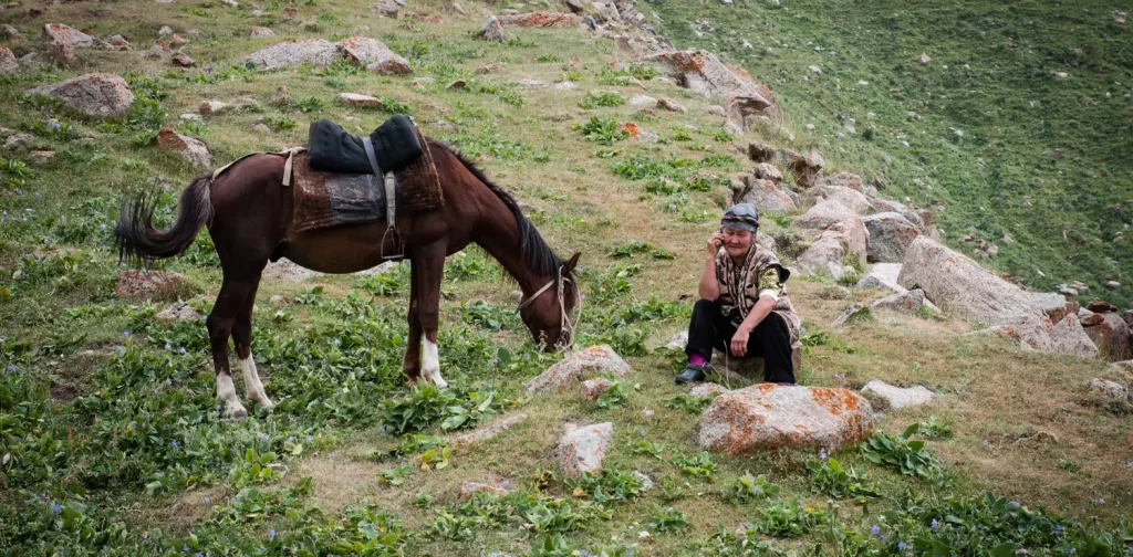 Seorang perempuan dewasa duduk di samping seekor kuda yang sedang makan di lereng gunung