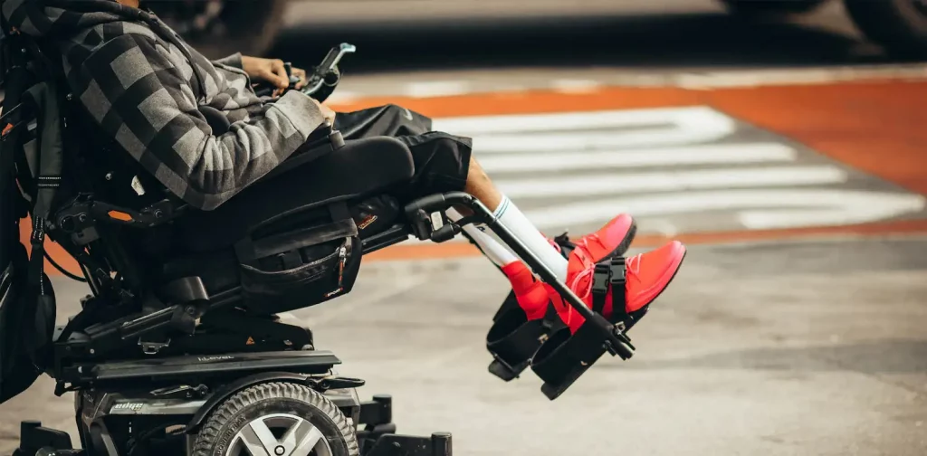 potret dari samping seseorang difabel yang mengenakan sepatu merah di kursi roda bermotor di pinggir jalan