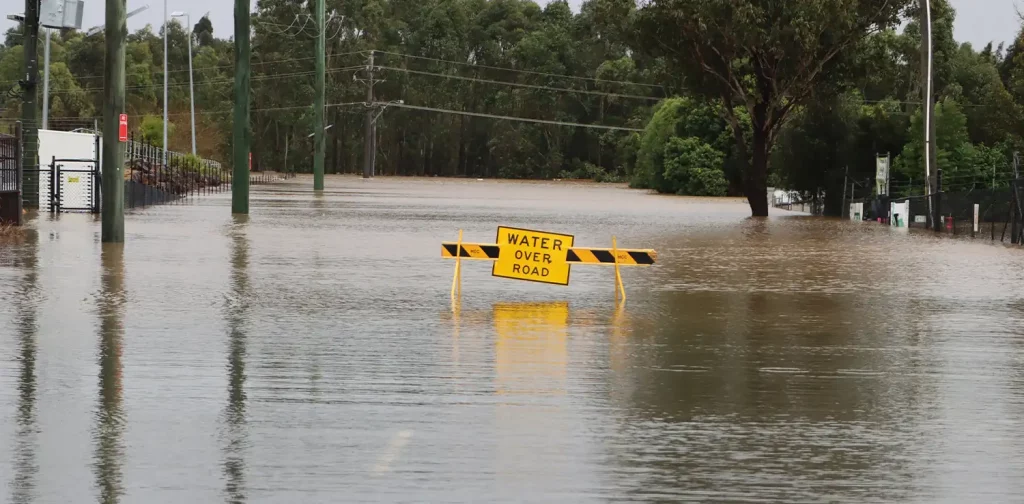 tanda peringatan berwarna kuning bertuliskan “Water Over Road” di tengah jalan yang banjir.