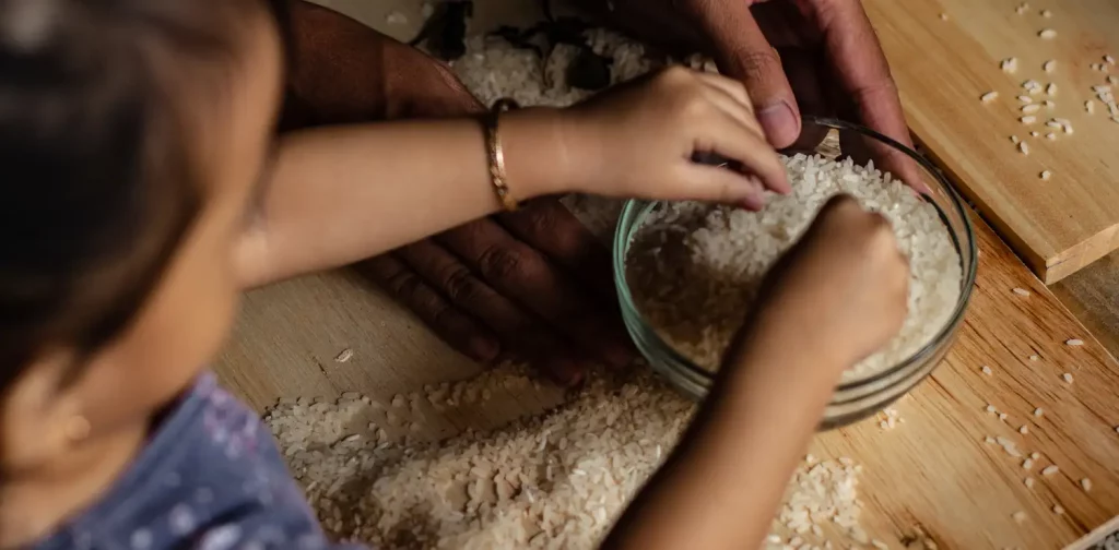 potret dari belakang bahu, seorang anak dengan rasa ingin tahu menyentuh semangkuk beras, dengan sepasang tangan orang dewasa memegang mangkuk