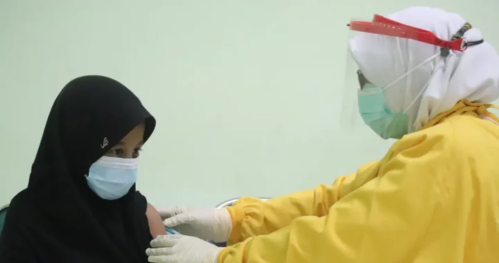 petugas kesehatan berbaju kuning dan memakai face-shield memegang lengan seorang pasien, bersiap-siap untuk menyuntikkan vaksin.