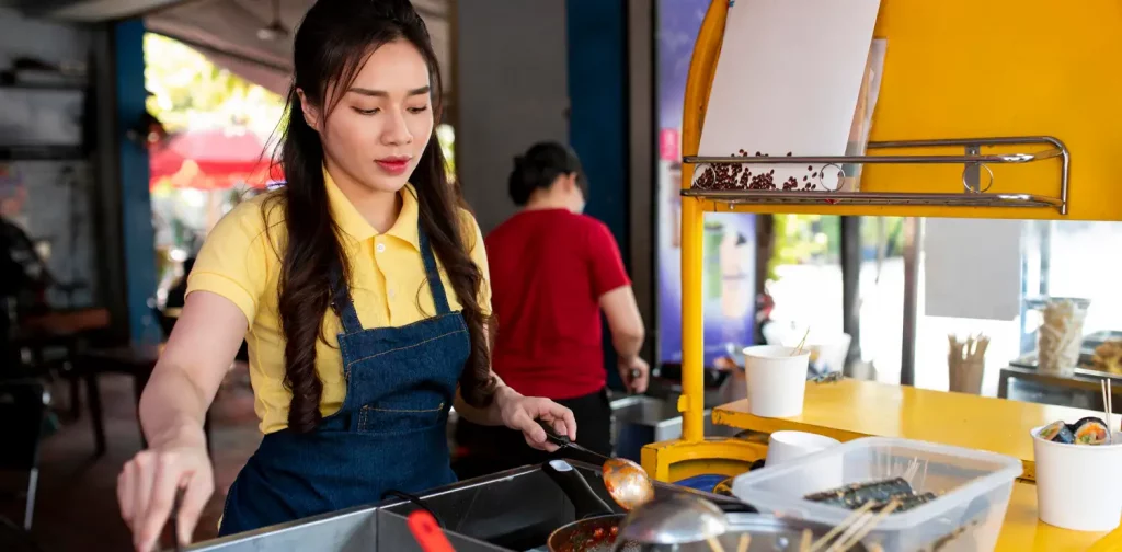 Seorang perempuan memakai overall biru dan baju kuning sedang menggoreng sesuatu di penggorengan.