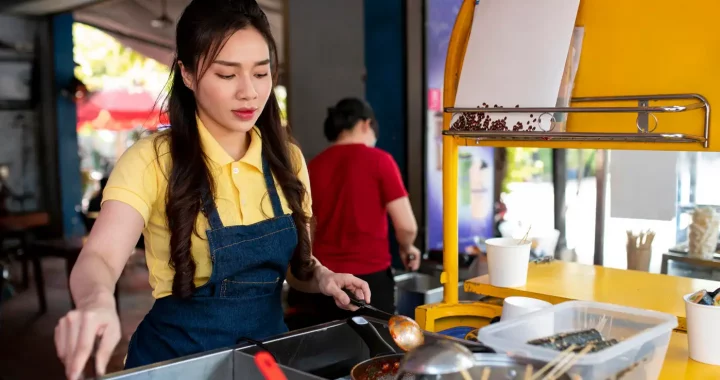 Seorang perempuan memakai overall biru dan baju kuning sedang menggoreng sesuatu di penggorengan.