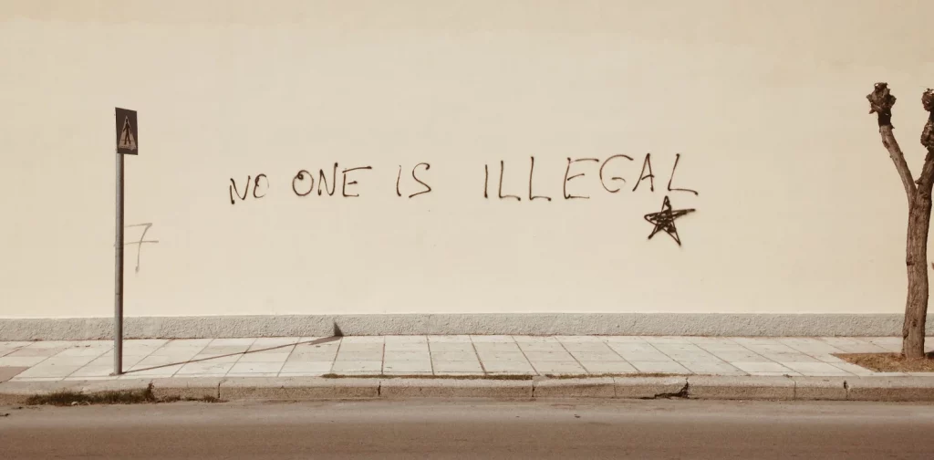 tulisan “No One is Illegal” ditulis di dinding putih di trotoar