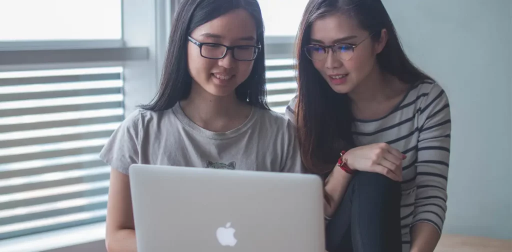 Dua perempuan tersenyum menatap layar laptop MacBook.