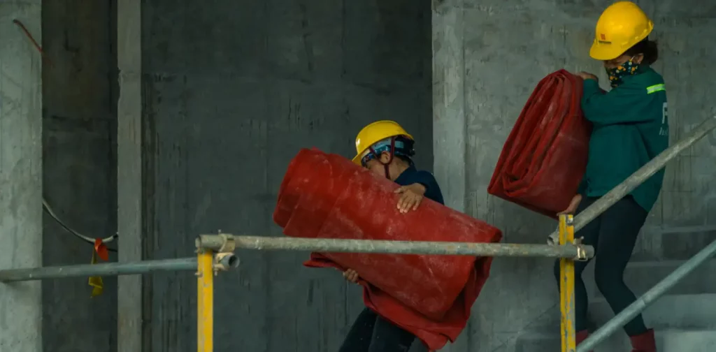 dua pekerja bertopi kuning mengangkat kantung plastik merah berukuran besar