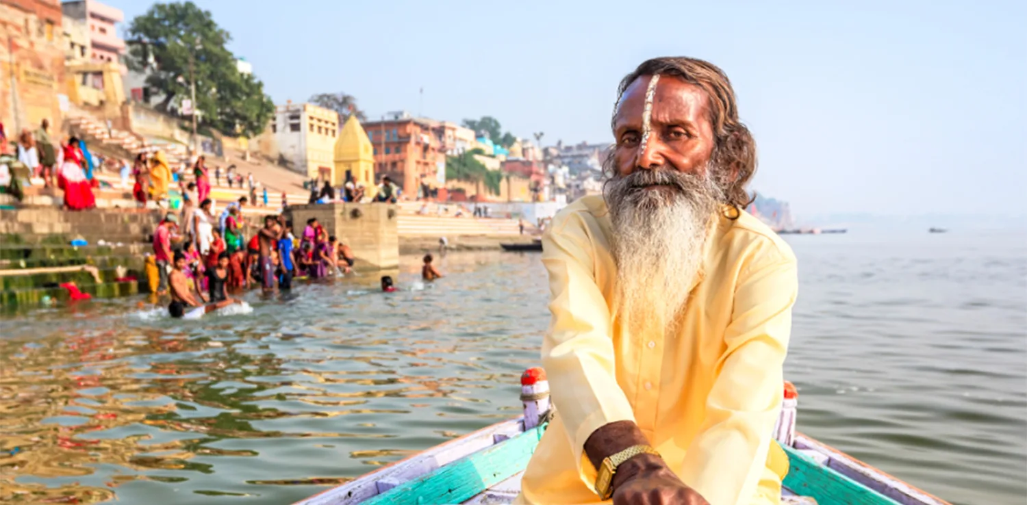 Seorang pria India mendayung perahu di cekungan Gangga, India, dengan latar belakang rumah-rumah dan orang-orang di tepi sungai.