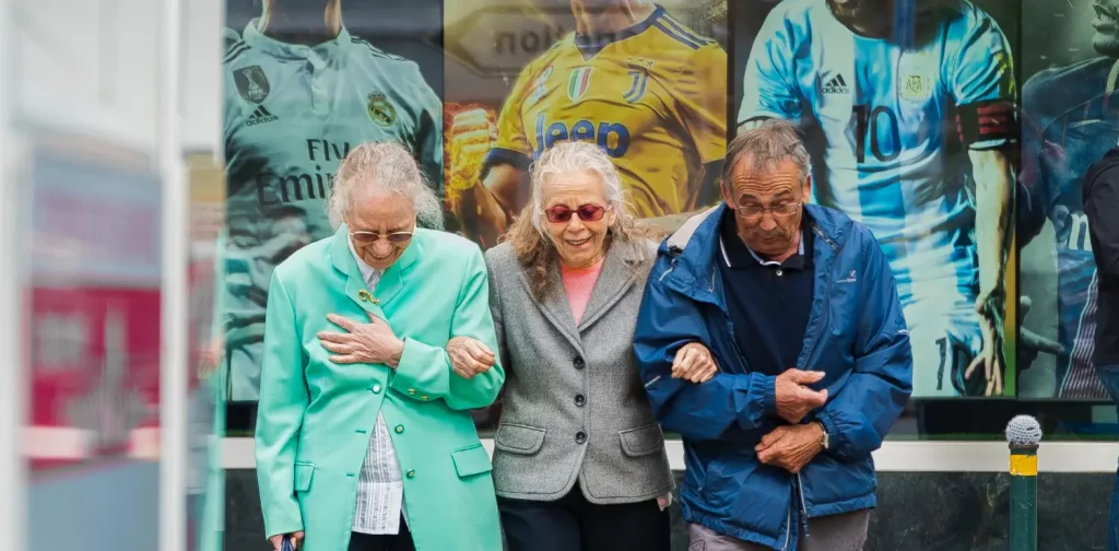 dua lansia perempuan dan seorang lansia laki-laki saling bergandengan tangan untuk menyeberang jalan