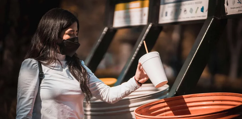 seorang perempuan bermasker memasukkan cangkir kosong ke dalam tong sampah
