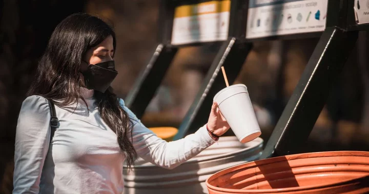 seorang perempuan bermasker memasukkan cangkir kosong ke dalam tong sampah