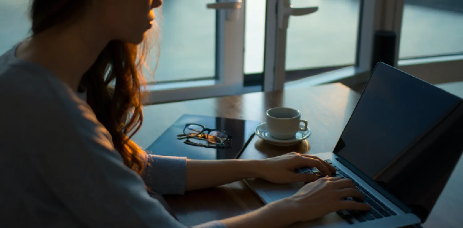 seorang perempuan pekerja duduk di hadapan laptop dengan layar gelap, dengan secangkir minuman dan kacamata di sampingnya.
