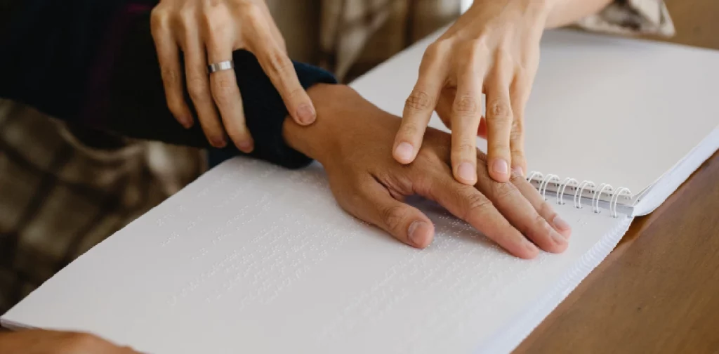 sepasang tangan memegang sebuah tangan di atas kertas bertuliskan huruf-huruf Braille.