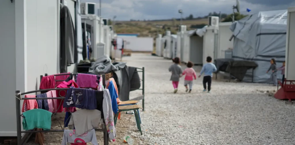 kamp pengungsi dengan latar tiga anak, pakaian tergantung, dan tenda.