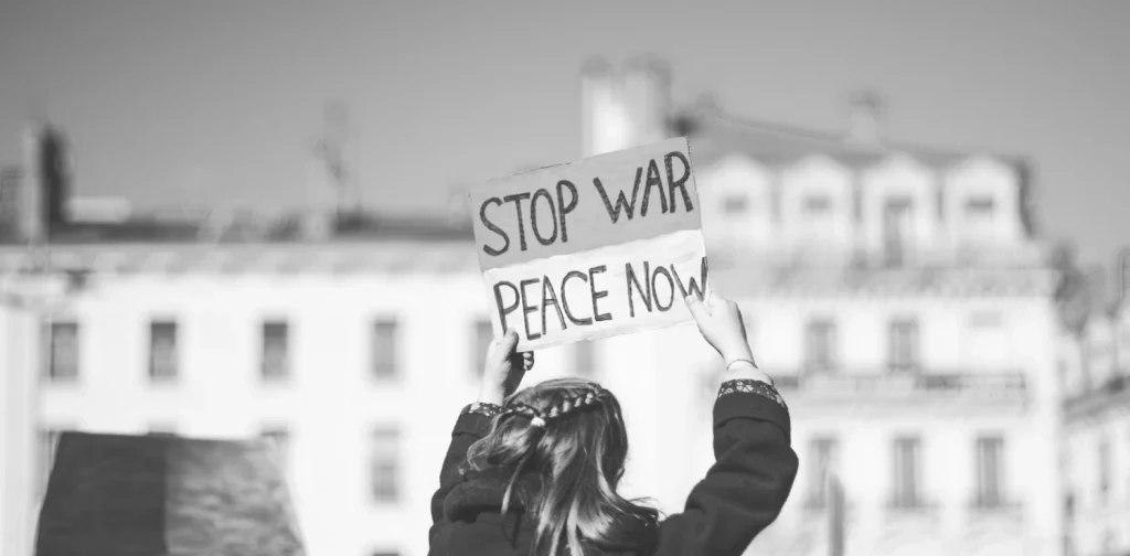 seorang perempuan mengangkat poster bertuliskan “Stop War, Peace Now” dengan latar bangunan bertingkat.