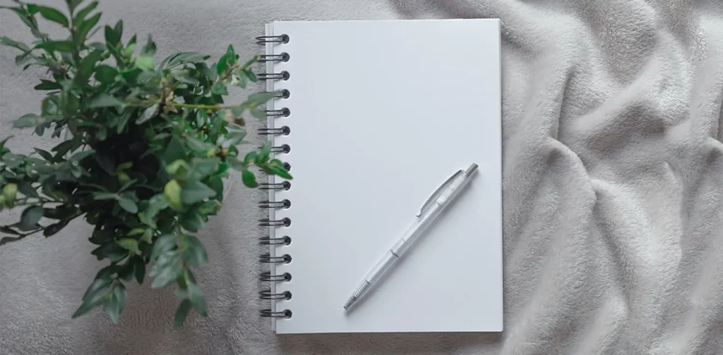 buku catatan kosong dengan pulpen putih dan tanaman hijau di atas meja berkain