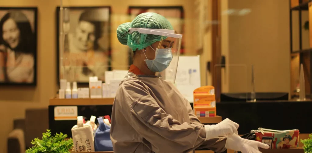 Seorang tenaga kesehatan perempuan dengan penutup wajah dan masker memakai sarung tangan medis.