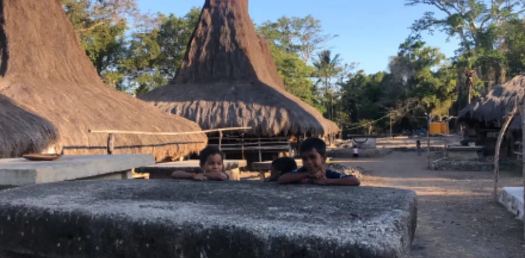 Tiga anak laki-laki berdiri di samping batu di dekat rumah adat Sumba di Kampung Kadoku
