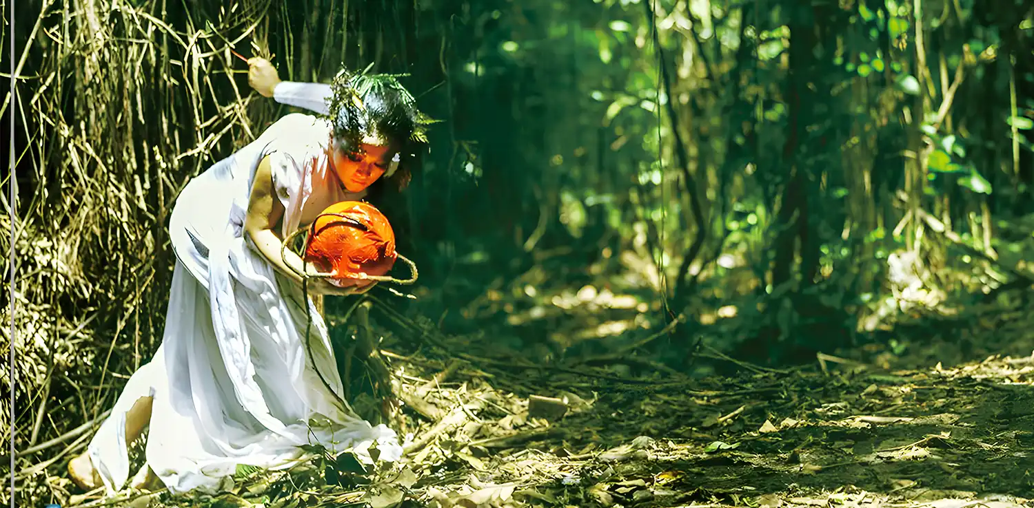 seorang perempuan menari di hutan dengan gaun putih dan memegang properti berupa bola berwarna oranye.