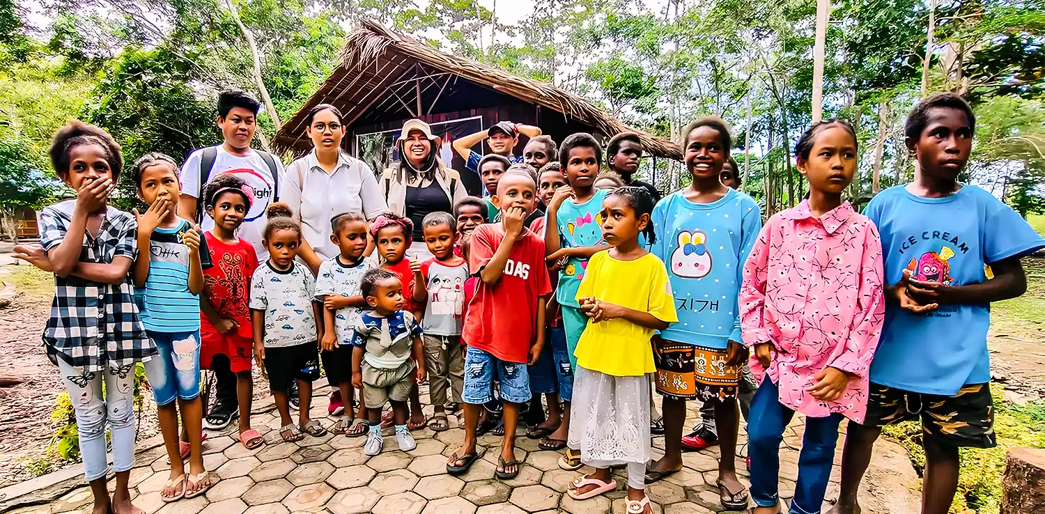 anak-anak Papua berfoto bersama beberapa sejumlah orang dewasa dengan latar bangunan beratap rumbia di belakang.