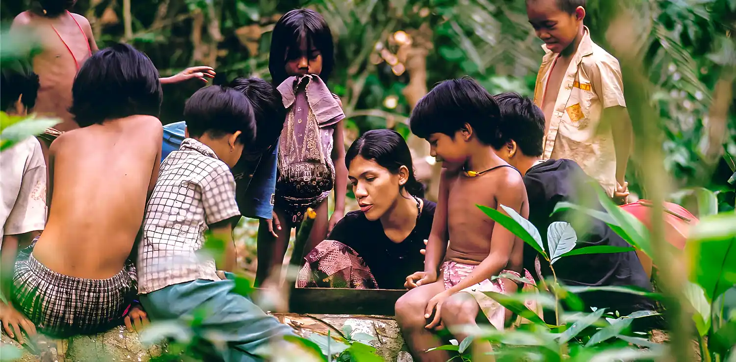 seorang perempuan sedang mengajar sesuatu di tengah hutan dikelilingi oleh anak-anak kecil.