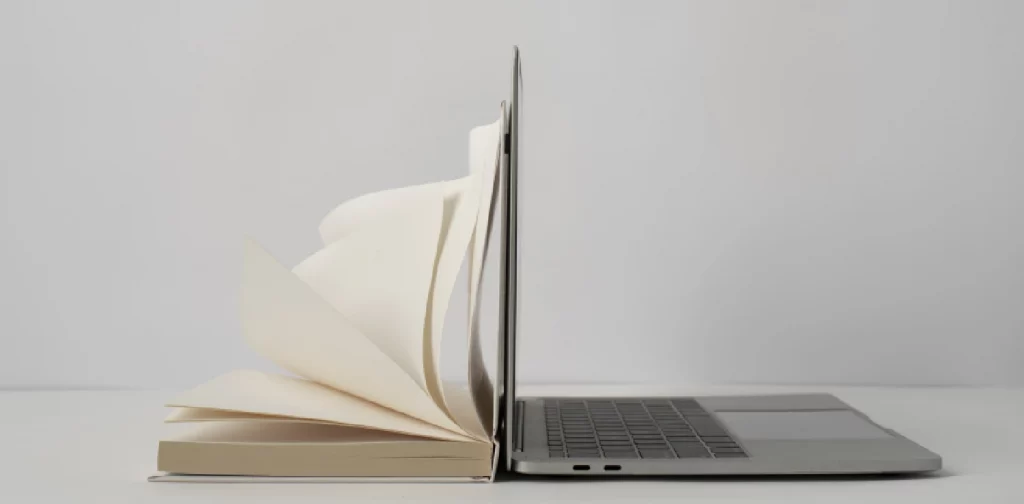 sebuah buku yang terbuka ditempatkan berdampingan dengan laptop yang terbuka