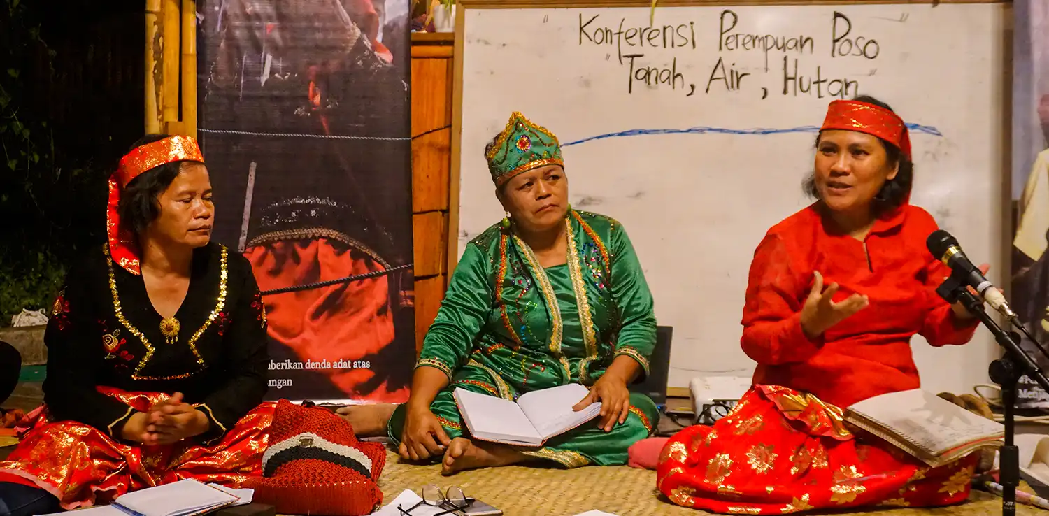 tiga perempuan berpakaian adat dengan ikat di kepala duduk lesehan dengan sebuah papan tulis di belakang bertuliskan ‘Konferensi Perempuan Poso: Tanah, Air, Hutan.