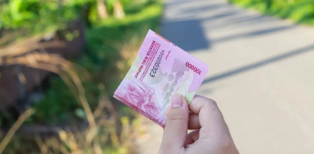 sebuah tangan memegang uang kertas seratus ribu rupiah dengan latar jalan dan pepohonan hijau.