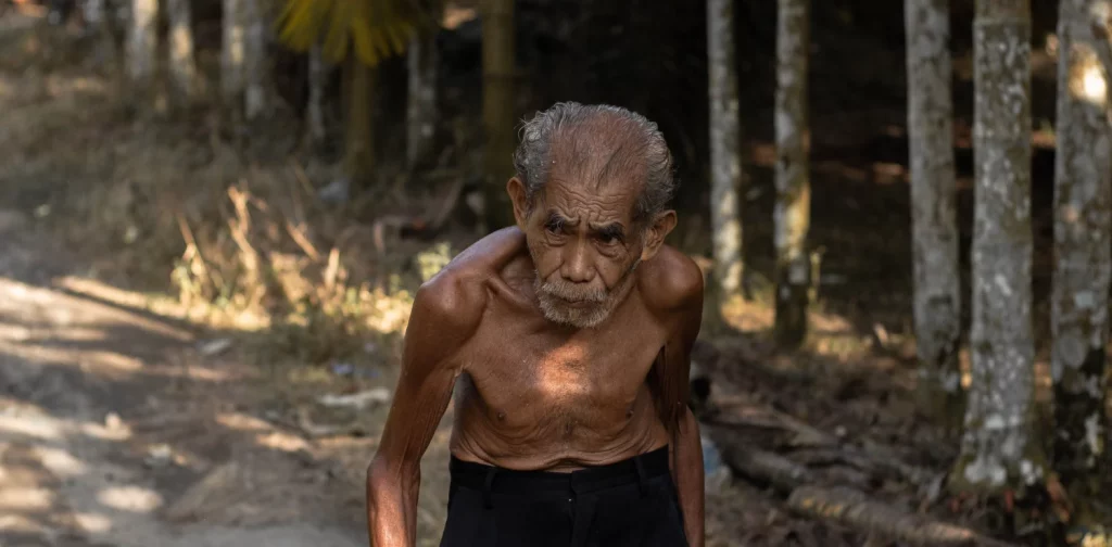 seorang pria tua tanpa baju berjalan dengan badan membungkuk dengan layar pohon pinang.