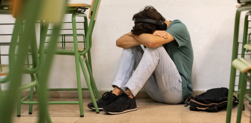 seorang remaja berjins biru dan berkaos hijau duduk di lantai di dekat kursi kelas dengan menekuk lutut dan membenamkan wajah di tangannya yang terlipat.