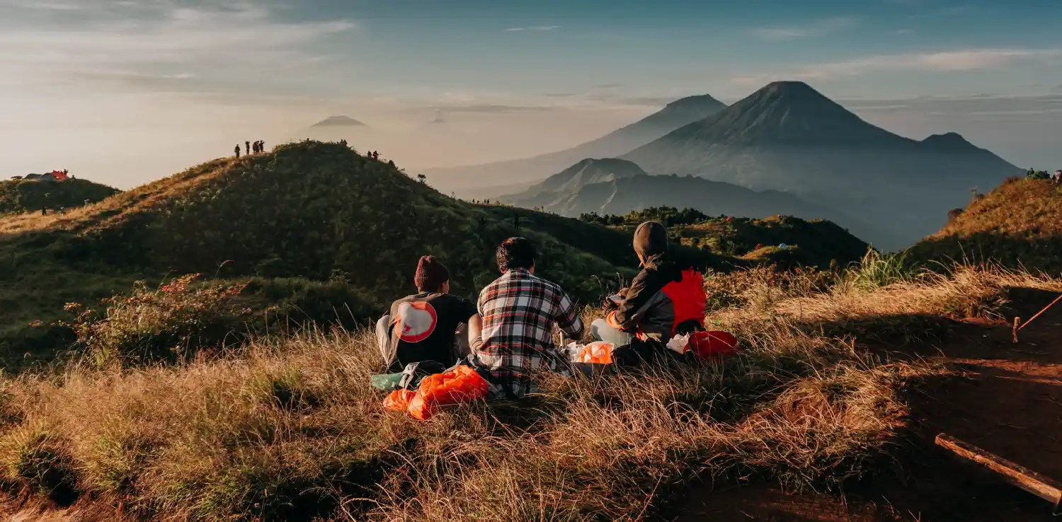 tiga orang duduk di rerumputan di puncak gunung dengan latar pegunungan di kejauhan.