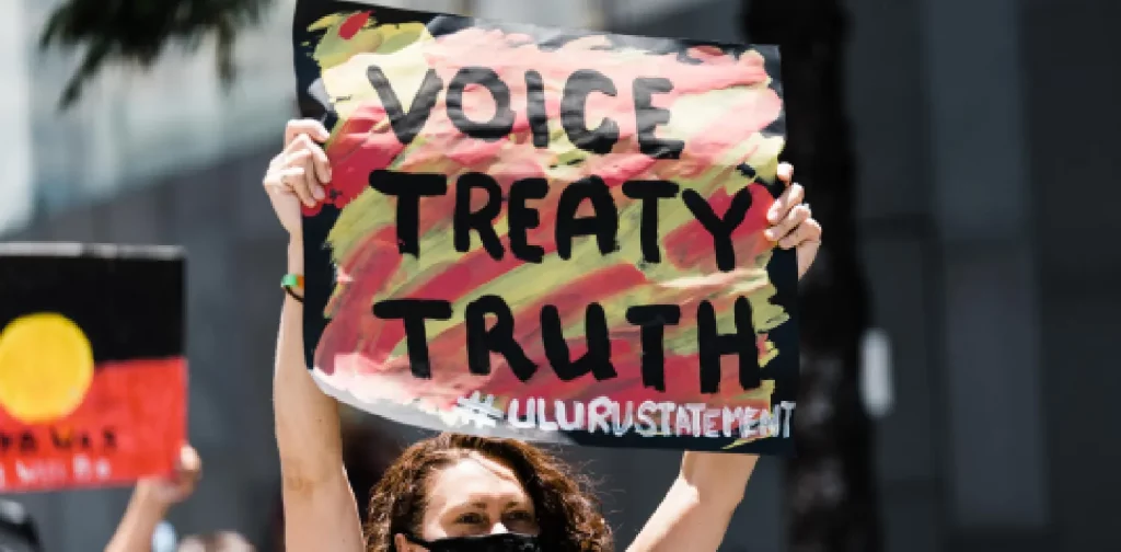 seorang perempuan bagian dari Masyarakat Adat Australia menjunjung poster bertuliskan “Voice Treaty Truth”