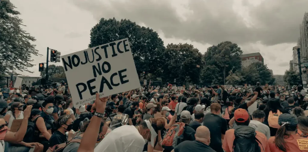 seseorang yang mengacungkan poster bertuliskan “No Justice, No Peace” di tengah kerumunan