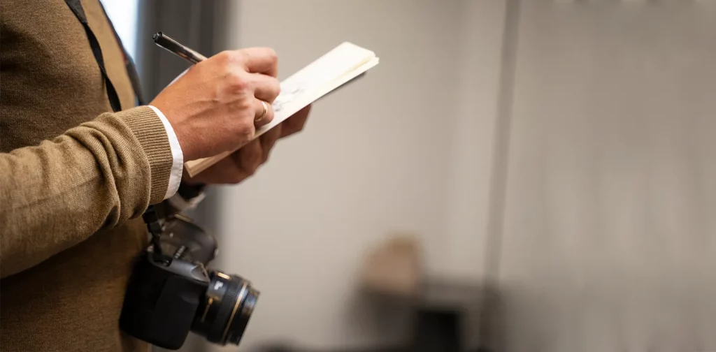 foto tangan memegang bolpen dan notes dengan kamera yang tergantung di dada