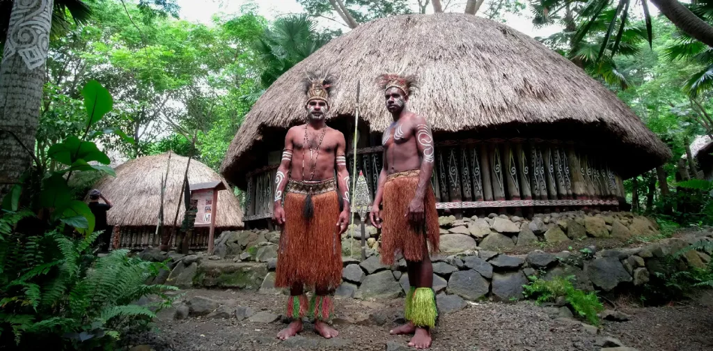 dua lelaki berpakaian adat berdiri di depan rumah adat honai