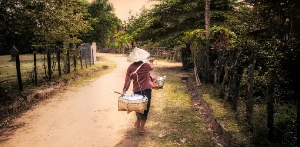 seorang perempuan berjalan di jalanan tanah sambil membopong dua keranjang dengan kayu