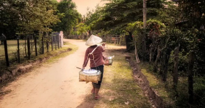 seorang perempuan berjalan di jalanan tanah sambil membopong dua keranjang dengan kayu