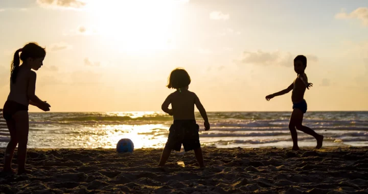 Tiga orang anak sedang bermain bola di pantai.