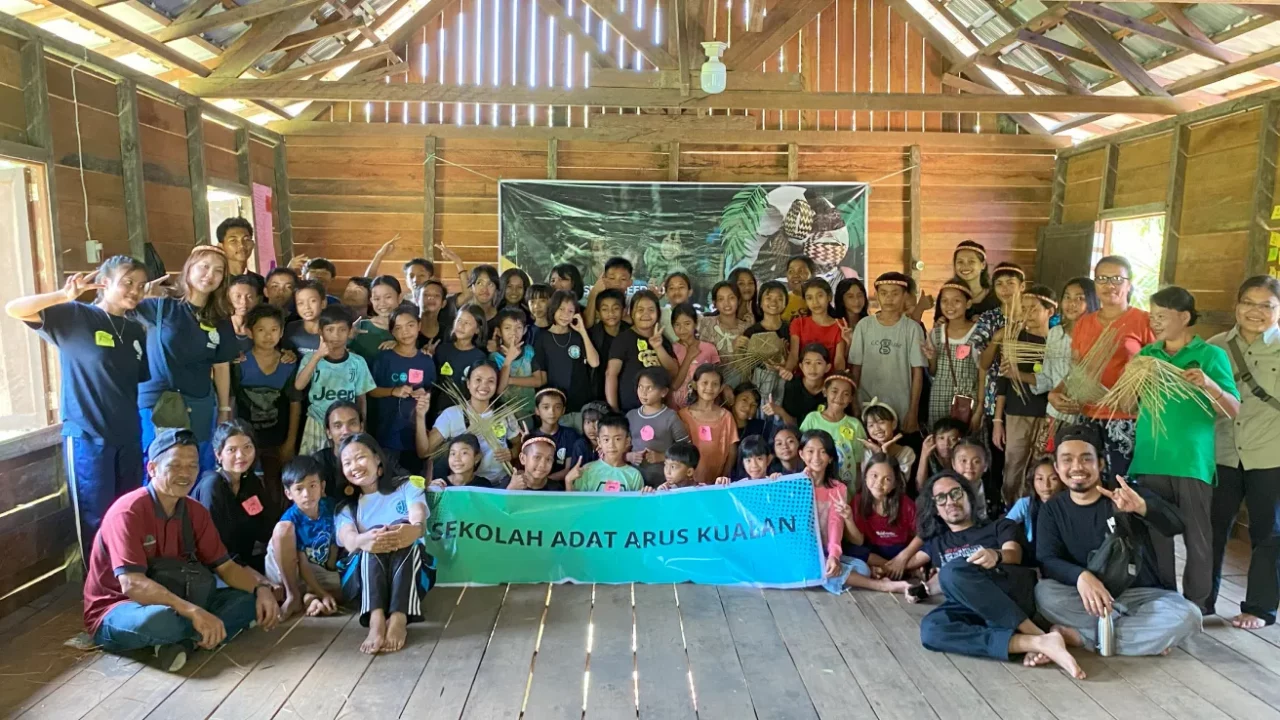 sekelompok anak-anak dan pemuda berfoto bersama dengan spanduk bertuliskan SEKOLAH ADAT ARUS KUALAN.