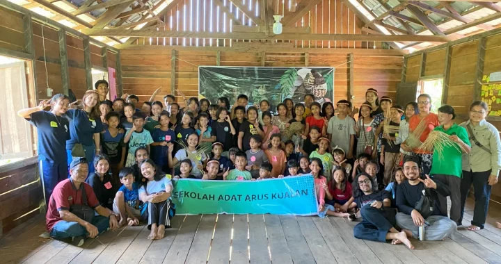 sekelompok anak-anak dan pemuda berfoto bersama dengan spanduk bertuliskan SEKOLAH ADAT ARUS KUALAN.