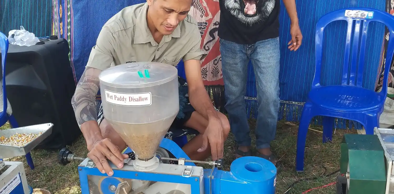 Dua orang laki-laki sedang berdiri di sebelah mesin penggiling padi bertenaga surya.