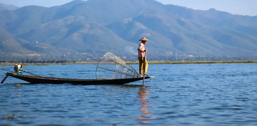 seseorang memakai caping berdiri di pinggir perahu di tengah danau saat siang hari