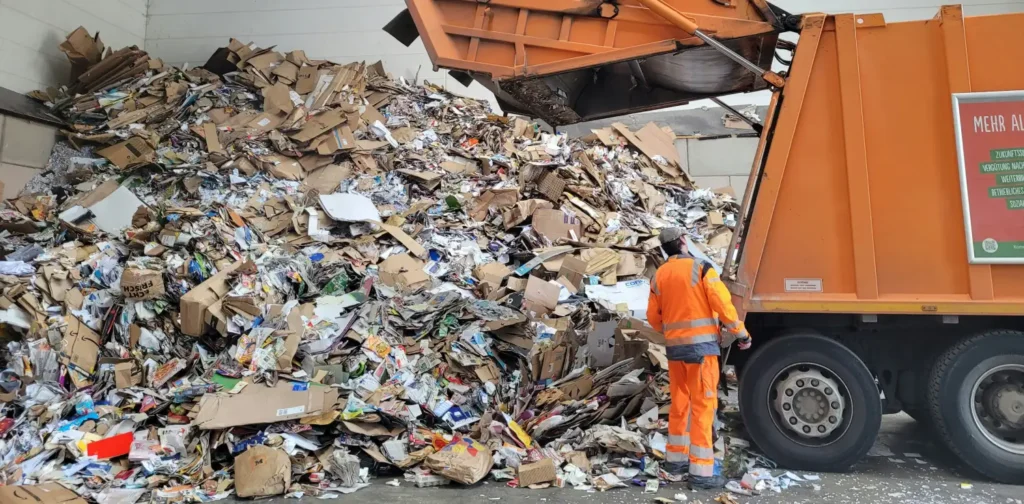 Seorang pria berpakaian oranye berdiri di samping tumpukan sampah dan truk pengangkut sampah