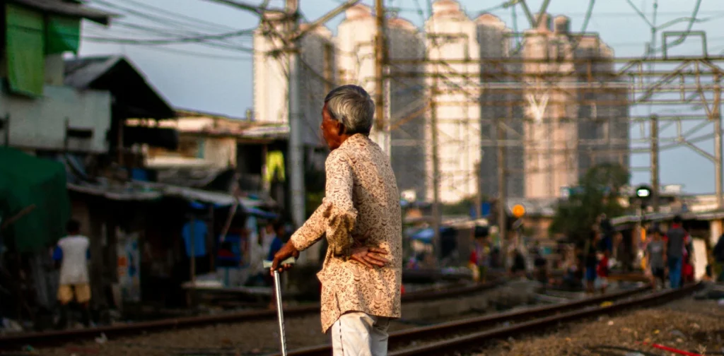 seorang pria tua berdiri di dekat rel kereta api dengan latar rumah-rumah gubuk