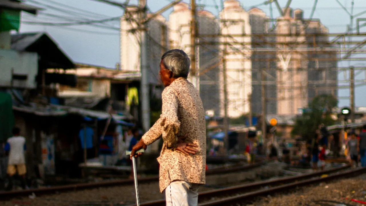 seorang pria tua berdiri di dekat rel kereta api dengan latar rumah-rumah gubuk